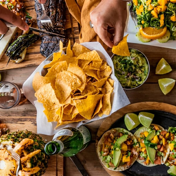 Overhead view of a wooden table laden with various Mexican dishes, drinks, and hands reaching for food.