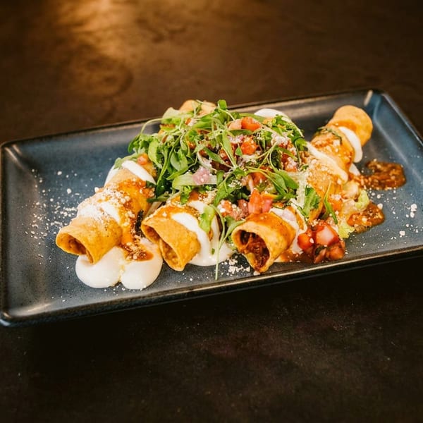 Four taquitos topped with sour cream, salsa, and fresh greens on a dark rectangular plate.