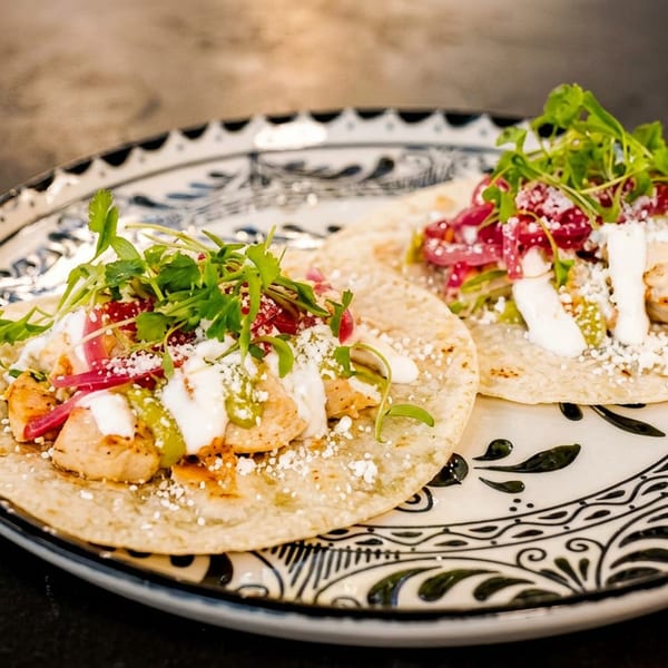 Two chicken tacos with red onions, cilantro, and cheese on a decorative plate.